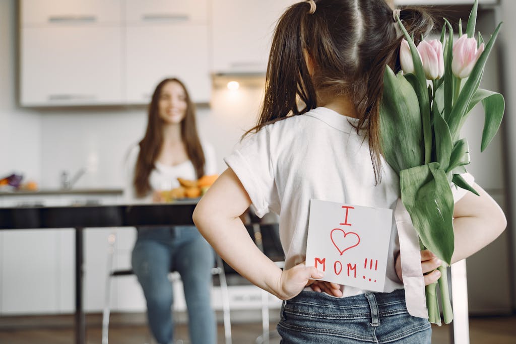 Back view of little girl with ponytails in casual clothes standing against mother with hands behind back and holding bouquet of tulips and drawing I love mom on kitchen